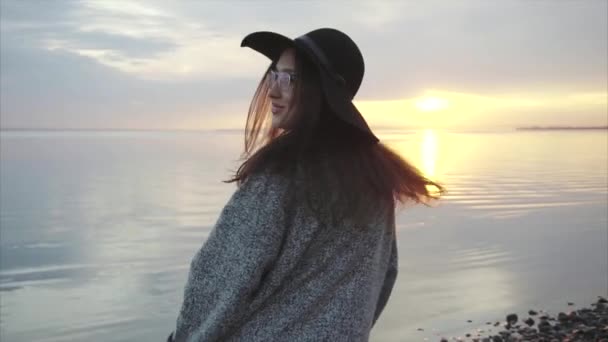 Jeune jolie femme en chapeau noir et lunettes près de la mer au coucher du soleil — Video