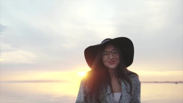 Jonge mooie lachende vrouw in zwarte hoed en bril in de buurt van de zee bij zonsondergang — Stockvideo