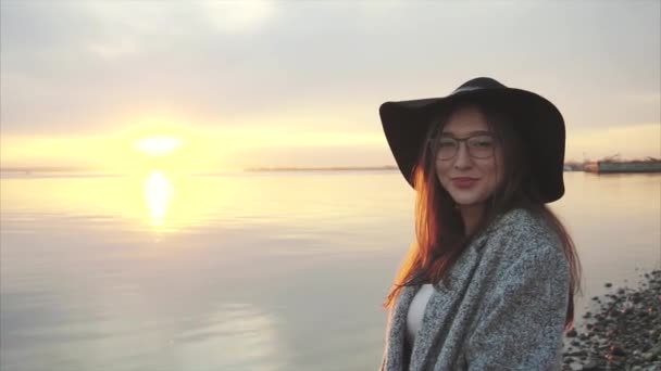 Joven mujer bastante sonriente en sombrero negro y gafas cerca del mar al atardecer — Vídeo de stock