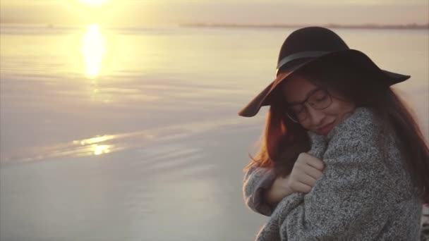 Jovem mulher bonita em chapéu preto e óculos perto do mar ao pôr do sol — Vídeo de Stock