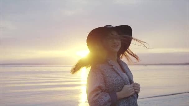 Jonge mooie en lachende vrouw in zwarte hoed en bril in de buurt van de zee bij zonsondergang — Stockvideo