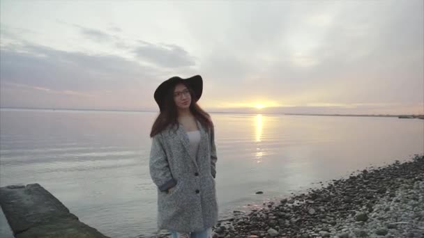 Young pretty and smiling woman in black hat and glasses near the sea at sunset — Stock Video