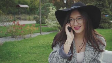 Genç kadın siyah şapka ve gözlük telefonda konuşurken parktaki bankta oturur