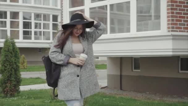 Красивая женщина в черной шляпе и в очках с чашкой кофе, идущая по улице — стоковое видео
