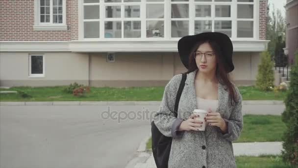Красивая женщина в черной шляпе и в очках с чашкой кофе, идущая по улице — стоковое видео