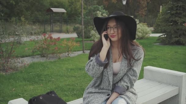 Mujer joven sentada en un banco en el parque de la ciudad y hablando por teléfono — Vídeos de Stock
