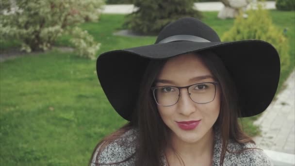 Närbild porträtt av ganska leende kvinna i svart hatt och glasögon i parken — Stockvideo