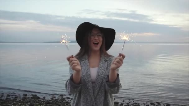 Mujer bonita saltando y regocijándose con chispas en la playa al atardecer — Vídeos de Stock