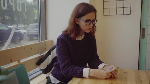 Chica en gafas sentado en la cafetería y navegar por Internet en el teléfono inteligente — Vídeos de Stock