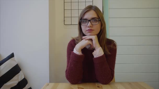 Nahaufnahme einer jungen brünetten Frau mit Brille, die drinnen in die Kamera schaut — Stockvideo