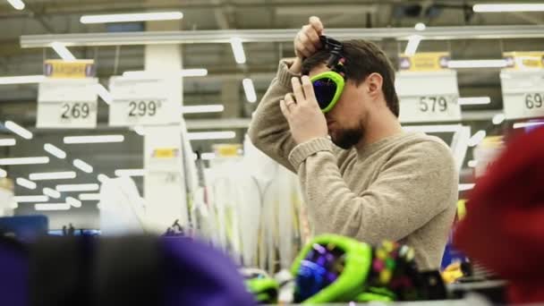 Junger Mann probiert im Supermarkt Snowboardbrille an. — Stockvideo