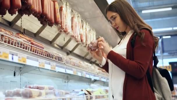 Mujer joven elige salchichas en el departamento de comestibles en el supermercado . — Vídeos de Stock