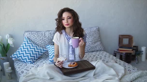Jonge vrouw ontbijten broodjes en thee in de slaapkamer op bed. — Stockvideo
