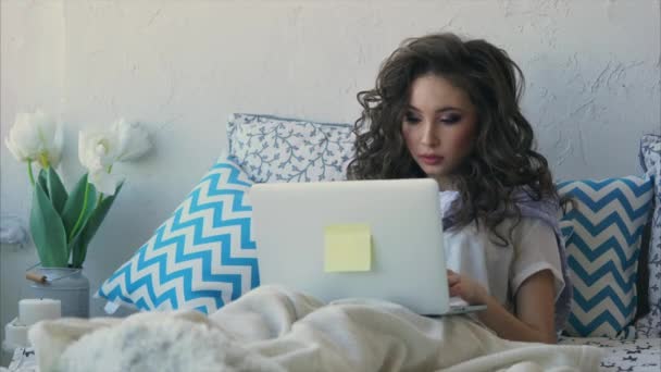 Young woman writes the text on a laptop in the bedroom, covered by a blanket. — Stock Video