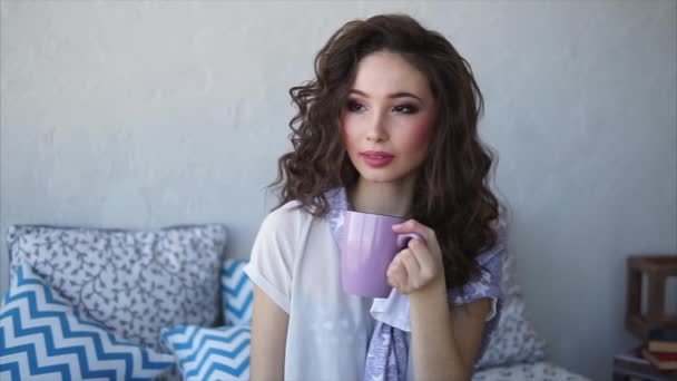 Mooie vrouw gekleed in pyjama's drinken koffie in de ochtend in haar slaapkamer. — Stockvideo