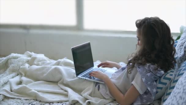 Ragazza adulta stampa testo sulla tastiera del computer portatile, sdraiato sotto una coperta a letto . — Video Stock