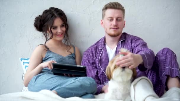 Schattig jong koppel verliefd zittend op een bed met een tablet en een hond aaien — Stockvideo