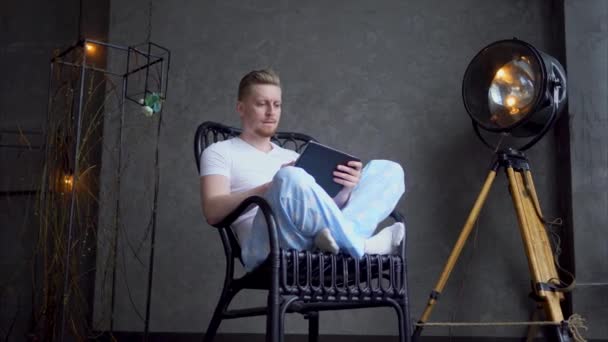 A young man who sits in a wooden chair works on a portable tablet. — Stock Video