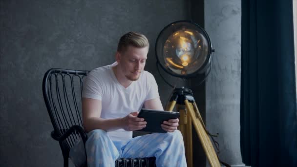 Mladý muž, oblečený v pyžamu, sedí v křesle pomocí přenosného tabletu. — Stock video