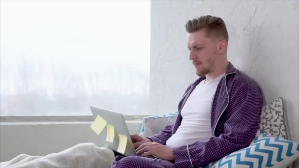 Homem com laptop sentado na cama se comunica com amigos em redes sociais . — Vídeo de Stock