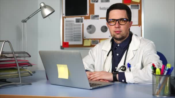 Doctor en bata blanca sentado en el gabinete y trabajando en su computadora portátil . — Vídeos de Stock