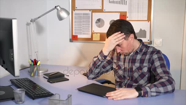 De man met een hoofdpijn zit achter het werk, op de tafel een heleboel apparaten — Stockvideo