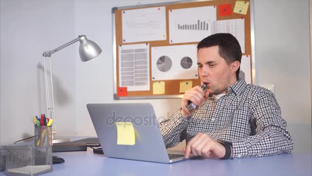 Jovem empresário no trabalho usando laptop. Ele vaping cigarro eletrônico . — Vídeo de Stock