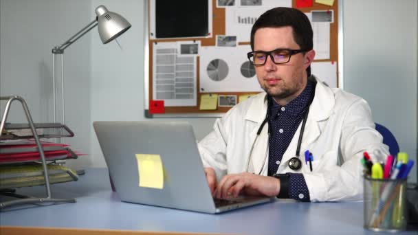 Doctor en bata blanca sentado en el gabinete y trabajando en su computadora portátil . — Vídeos de Stock