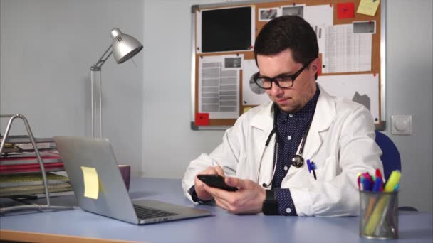 Αρσενικό γιατρό σε λευκό παλτό με στηθοσκόπιο κάθεται στο γραφείο και χρησιμοποιώντας έξυπνο ρολόι. — Αρχείο Βίντεο