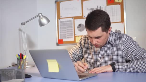 Αρσενικά χρησιμοποιούν φορητό υπολογιστή και παίρνει σημειώσεις σε χαρτί που κάθεται στο γραφείο του. — Αρχείο Βίντεο