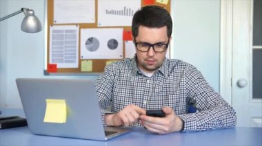 Dizüstü bilgisayar, o bilgileri bulmak için akıllı telefon kullanma ile ofiste çalışan adam.