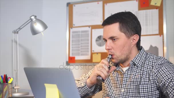 Человек курит электронную сигарету в своем кабинете, он смотрит на монитор ноутбука — стоковое видео