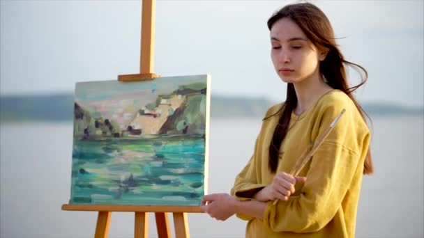 Романтичная девушка-художник стоит рядом с ее натюрмортом на берегу моря или реки — стоковое видео