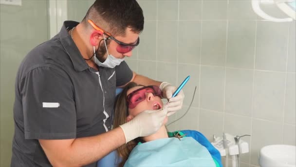 Médico ortodontista com óculos branqueia os dentes de uma jovem com um laser — Vídeo de Stock