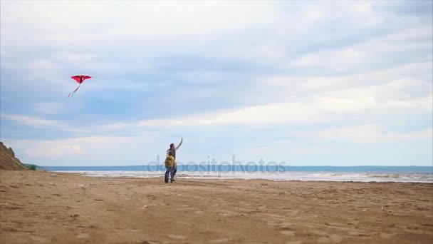 El joven padre y su hijo corren felices por la playa en clima frío con una cometa — Vídeo de stock