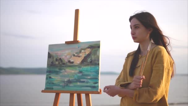 Romantic girl artist stands near her still life next to the sea or river — Stock Video