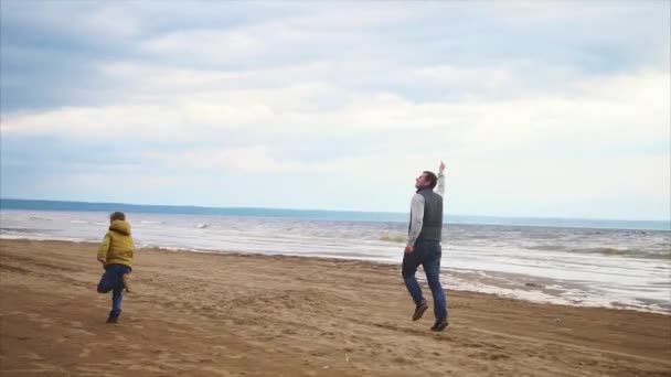 Junger Vater und sein Sohn laufen bei kaltem Wetter fröhlich mit einem Drachen am Strand entlang — Stockvideo