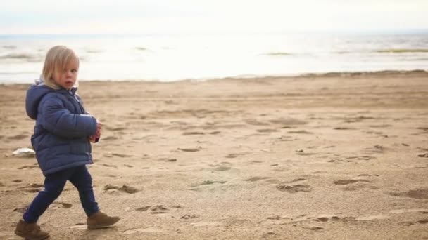 Маленький мальчик с светлыми волосами прогуливается вдоль песчаного пляжа у моря — стоковое видео