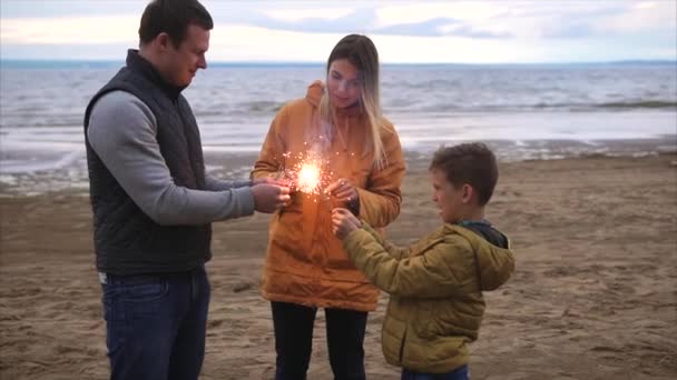 Familia joven encendiendo luces de bengala en una orilla de un río. Celebración familiar — Vídeos de Stock