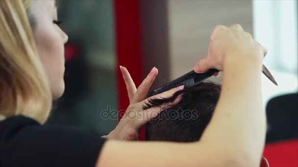 Mujer peluquera haciendo corte de pelo a un hombre joven. Ella usa tijeras y peine — Vídeo de stock