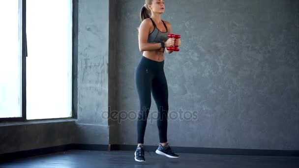 Joven mujer de fitness haciendo sentadilla con pesas en las manos. Escena en el gimnasio — Vídeo de stock