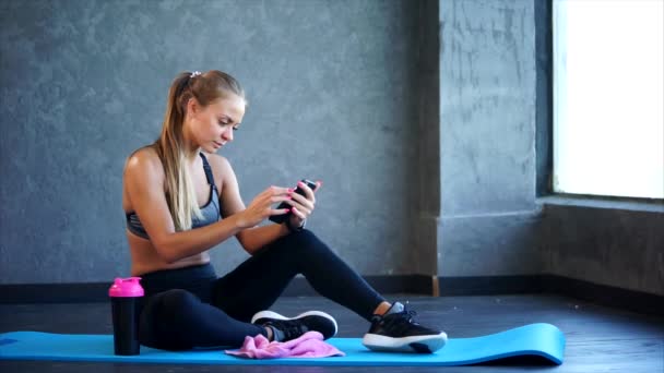 Mujer en el gimnasio con smartphone. Ella sentada en la alfombra y usando un teléfono inteligente — Vídeos de Stock