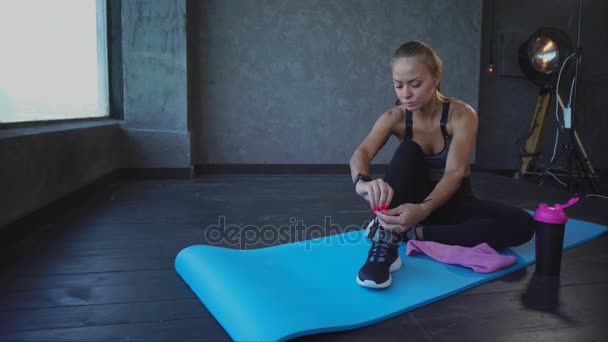 Молодой спортсмен завязывает шнурки на кроссовках, дама сидит на ковре для йоги — стоковое видео