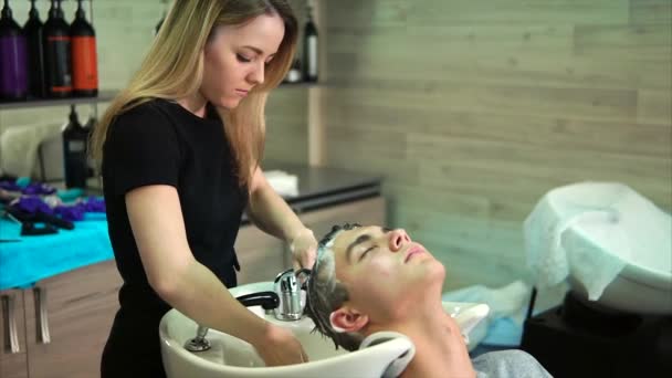 Um jovem cabeleireiro lava a cabeça do visitante de um salão de beleza com um xampu — Vídeo de Stock