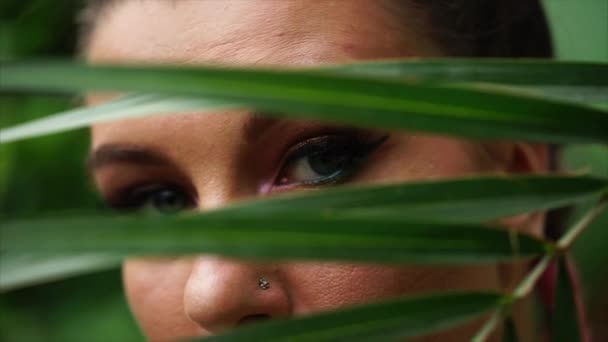 Крупним планом красива молода жінка дивиться за пальмовим листом . — стокове відео