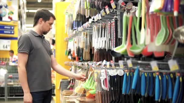 Um homem na loja perto do balcão de utensílios. Ele está à procura de espátula para sua esposa . — Vídeo de Stock
