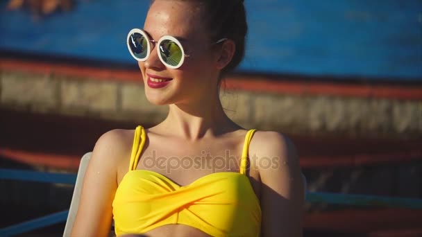 Κομψή γυναίκα σε ένα μαγιό και γυαλιά ηλίου, χαμόγελο, κάθεται έξω από το καλοκαίρι — Αρχείο Βίντεο