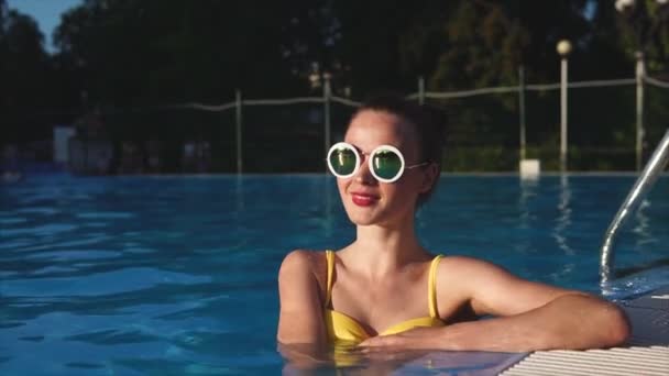 Een mooie vrouw in stijlvolle glazen is in het zwembad, ze houdt van een warme dag — Stockvideo