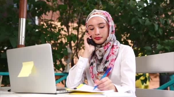 携帯電話で話していると、ノートブック内のノートを作る若いイスラム教徒の女性 — ストック動画