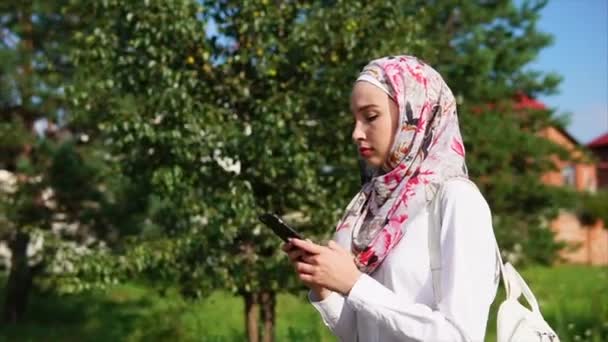 A young Muslim woman in a headscarf is chatting on a smartphone in the park — Stock Video